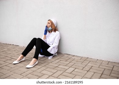 Dirty nurse with ash on face sitting outside hospital infirmary smoking cigarette after hard working day or surgery. Tired exhausted doctor woman dressed white medical gown have a rest due to stress

