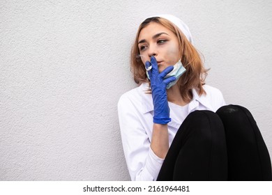 Dirty nurse with ash on face sitting outside hospital infirmary smoking cigarette after hard working day or surgery. Tired exhausted doctor woman dressed white medical gown have a rest due to stress

