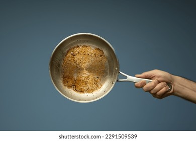 Dirty oily burnt metal frying pan held in hand by a male hand. Close up studio shot, isolated on a light blue background.