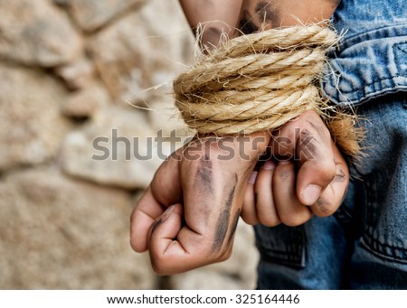 Dirty prisoner bound with rope. Hands behind back standing next to a wall.