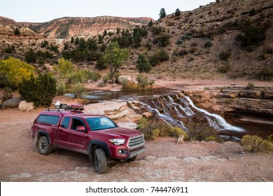 A Dirty Red Truck With Camper Shell, Propane Tank, Ammo Boxes, Gas Can, And Firewood On The Roof. The Truck Sits On A Rough, Rocky Trail Near A Waterfall On The Red Rock Sandstone Of Southern Utah.