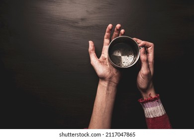Dirty, skinny child’s hands holding metal cup. Poverty and access to water concept. Black background, layout with free copy (text) space. Captured from above (top view).