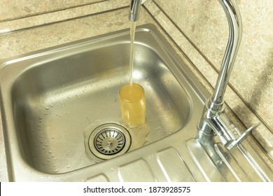 Dirty tap water flows out of the mixer. A glass tumbler filled with water stands in a stainless steel sink.