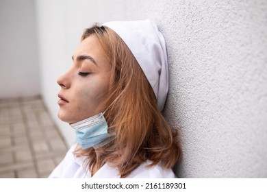 Dirty tired exhausted nurse with ash on face sitting outside hospital infirmary after hard working day or surgery. Doctor woman dressed white medical gown, face mask have a rest due to stress