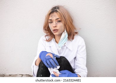Dirty tired exhausted nurse with ash on face sitting outside hospital infirmary after hard working day or surgery. Doctor woman dressed white medical gown, face mask have a rest due to stress
