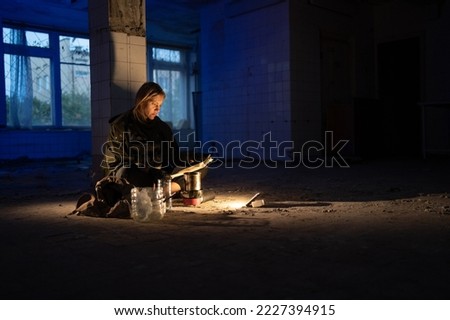 dirty tired woman sits on the floor in an abandoned room at night reads a newspaper shines with a lantern. Post-apocalypse. hiking in a post-apocalyptic world. copy space.