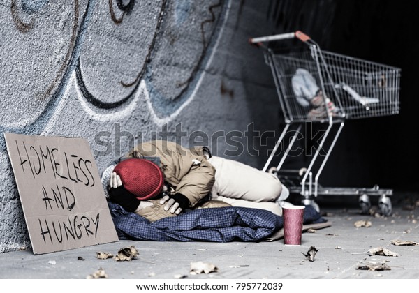 Dirty tramp lying on blanket next to a sign and\
trolley in the shelter of the\
city