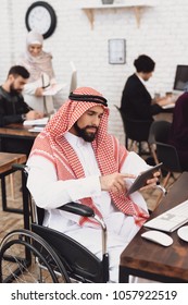 Disabled arab man in thawb in wheelchair working in office. Man is using tablet.