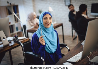 Disabled arab woman in hijab in wheelchair working in office. Woman is working on desktop computer.
