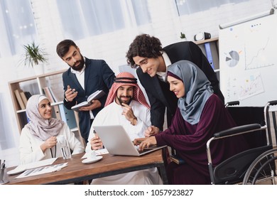 Disabled arab woman in hijab in wheelchair working in office. Woman is sharing notes on laptop.