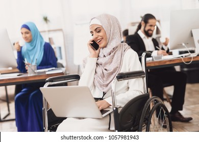 Disabled arab woman in hijab in wheelchair working in office. Woman is working on laptop and talking on phone.