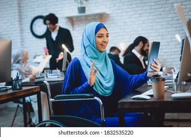 Disabled arab woman in hijab in wheelchair working in office. A woman sits and greets someone in a computer.