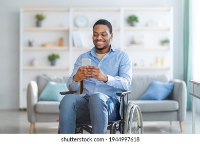 Disabled black man in wheelchair using smartphone, browsing web or watching movie at home. Handicapped young guy checking social media, speaking to friend on mobile device