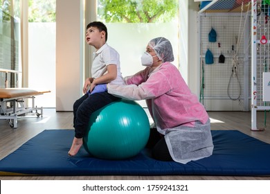 A Disabled Child Is On Top Of A Large Ball Doing Physical Therapy, Rehabilitation, Together With A Woman Who Is A Physiotherapist, Osteopath, Nurse. She Is Wearing A Pink And White Protective Su