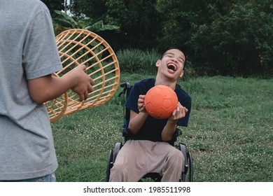 Disabled Child On Wheelchair Is Playing Basketball On The Lawn In Front Of The House Like Other People, Lifestyle Of Special Child,Life In The Education Age Of Children, Happy Disability Kid Concept.