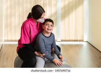 Disabled Child And Physiotherapist On Top Of A Peanut Gym Ball Doing Balance Exercises. Pandemic Mask Protection