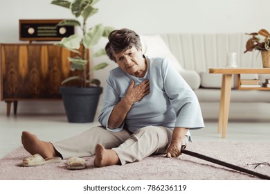 Disabled elder lady sitting on the floor with a walking stick and holding her chest after falling down