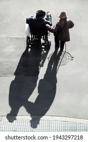 Disabled Elderly Man In Wheelchair Helped By His Partner Crossing The Street Road , Backlight With Shadows