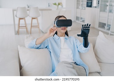 Disabled Girl With Cyber Arm In Vr Glasses. Handicapped Person Gets Rehabilitation At Home. Young Attractive European Woman Is Sitting In Living Room On Couch. Healthcare Innovation Technology.