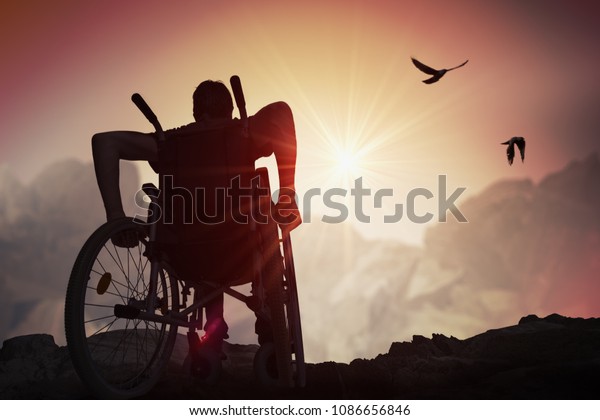 Disabled handicapped man has\
a hope. He is sitting on wheelchair and stretching hands at\
sunset.