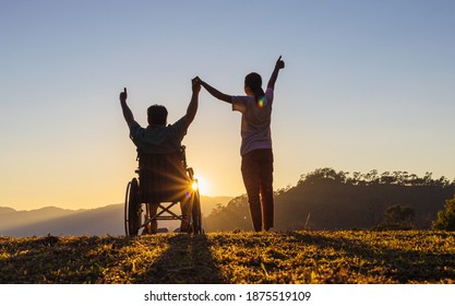 Disabled handicapped young man in wheelchair raised hands with his care helper in sunset.Silhouette