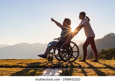 Disabled handicapped young man in wheelchair walking with his care helper in sunset.Silhouette