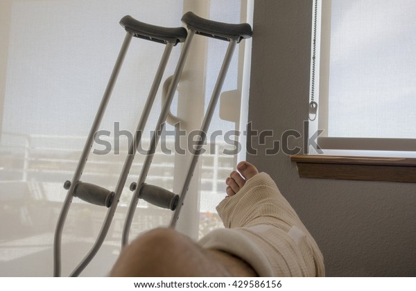 Disabled Injured Person With Sprained or\
Broken Ankle or Foot Sits Inside With Crutches Looking Outside the\
Sliding Glass Door Window on a Sunny\
Day.