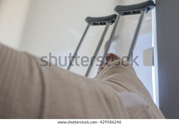 Disabled Injured Person With Sprained or\
Broken Ankle or Foot Sits Inside With Crutches Looking Outside the\
Sliding Glass Door Window on a Sunny\
Day.