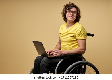 behinderter Mann sitzt in einem Rollstuhl mit Laptop und arbeitet online. Kurz junges Männchen auf beigem Hintergrund mit Lächeln auf Kamera einzeln auf beigem Hintergrund