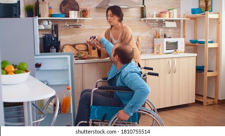 Disabled Man In Wheelchair Helping Wife In Kitchen. Opening Refrigerator. Guy With Paralysis Handicap Disability Handicapped Difficulties Getting Help For Mobility From Love And Relationship