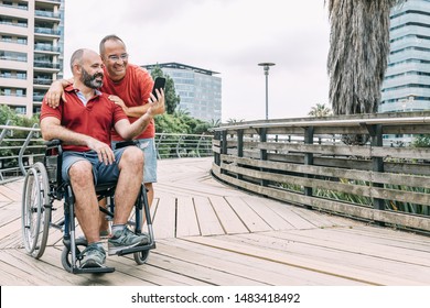 Disabled Man In Wheelchair Making A Selfie With His Phone With A Friend During A Walk, Concept Of Friendship And Integration Of People With Disabilities And Reduced Mobility Problems