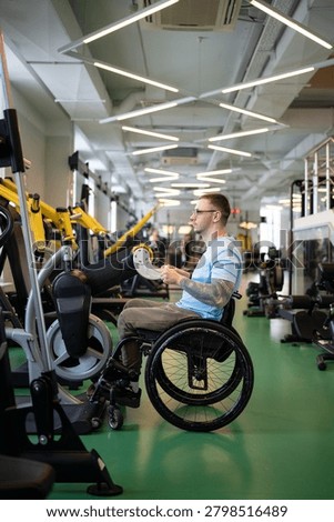 Disabled man in wheelchair using upper body ergometer for recovery after spinal injury, hand cycling machine helps build muscles strength. Male with spinal injury undergoing long-term rehabilitation.