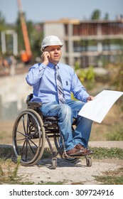 Disabled Person In The Construction Helmet With Documents In Hand Talking On The Phone On The Background Of Building. Successful Wheelchair User Directs The Work.