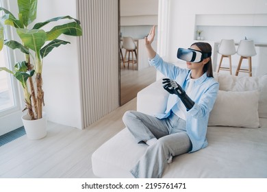 Disabled Person Gets Rehabilitation And Playing In Cyber Space. Young Caucasian Woman Sitting In Living Room On Couch. Handicapped Girl In Vr Glasses. Amputee With Bionic Limb Prosthesis.