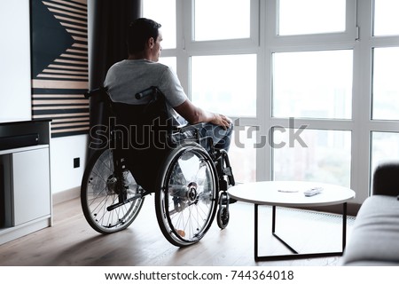 The disabled person in a wheelchair sits in front of a large panoramic window and looks into it. He is sad. He is in the living room of his modern apartment.
