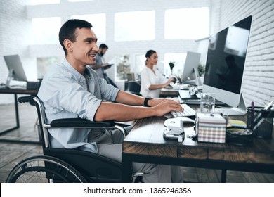 Disabled Person In The Wheelchair Works In The Office At The Computer. He Is Smiling And Passionate About The Workflow. Office People Working Together.