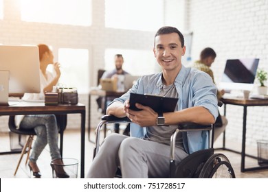 Disabled Person In The Wheelchair Works In The Office. He Writes In A Tablet For Papers. Man Is Smiling. His Colleagues Work Nearby.