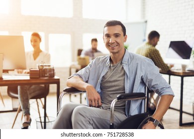 Disabled Person In The Wheelchair Works In The Office. He Is Posing For The Camera. Man Is Smiling. His Colleagues Work Nearby.