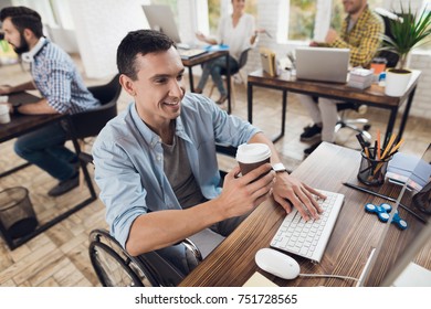 Disabled Person In The Wheelchair Works In The Office At The Computer. He Is Smiling And Passionate About The Workflow. Man Is Holding A Cup Of Coffee.