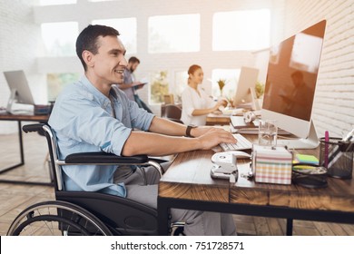 Disabled Person In The Wheelchair Works In The Office At The Computer. He Is Smiling And Passionate About The Workflow.