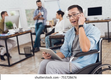 Disabled Person In The Wheelchair Works In The Office. He Works With Glasses On The Tablet. His Colleagues Work Nearby. Man In Wheelchair Is Talking On A Phone.