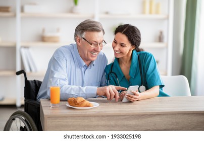 Disabled Senior Man And Young Nurse Using Smartphone Together, Browsing Web Or Watching Movie Indoors. Handicapped Elderly Patient And His Caregiver Checking Social Media On Mobile Device