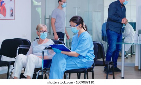 Disabled senior woman in wheelchair talking with nurse that wears protection mask against infection with coronavirus. Patient and medical staff in waiting area. Doctor in examination room.