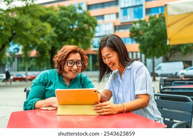 Disabled woman in electric wheelchair and chinese female friend using tablet sitting on sidewalk cafeteria - Powered by Shutterstock