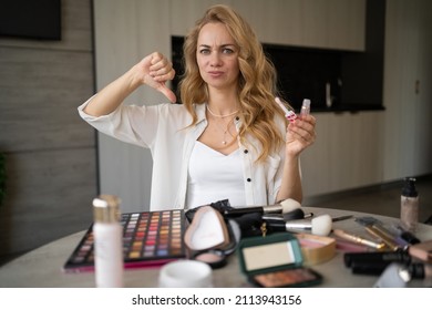 Disagreeing young attractive caucasian ethnic model vlogging on smartphone on tripod and showing lipstick and thumb down at table with makeup cosmetics. Bad review and unhappy customer. portrait