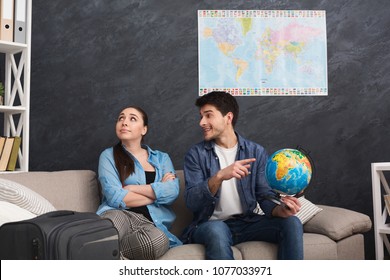 Disagreement In Choosing Place Of Vacation. Couple Looking For Compromise, Man Pointing On Place On Globe, Sitting With Stuff At Home, Copy Space