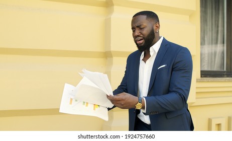 Disappointed African Businessman Walking With Papers On Urban Street. African American Business Man Reading Documents Outdoors. Confused Afro Male Professional Examining Paperwork Outside.