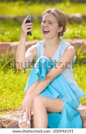 Disappointed and Angry Young Caucasian Woman Shouting at Her Cellphone. Sitting on the Ground Against Green Grass. Vertical Image Composition