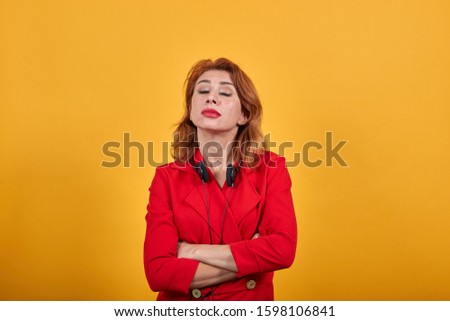 Disappointed caucasian young woman having nice headphones, keeping hands crossed with closed eyes wearing fashion red jacket over isolated orange background. People lifestyle concept.