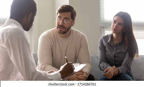 Disappointed Couple At Reception With Family Therapist. African American Doctor Listening Man In Bad Mood. Family Relationship Disorder Concept. Psychology Session With Coach